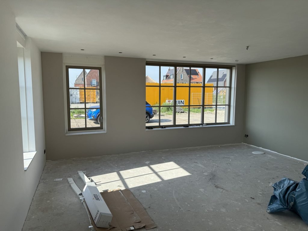 Ruime kamer in een nieuw huis met warm grijze wanden, sprossenramen en zonlicht op de kale vloer
