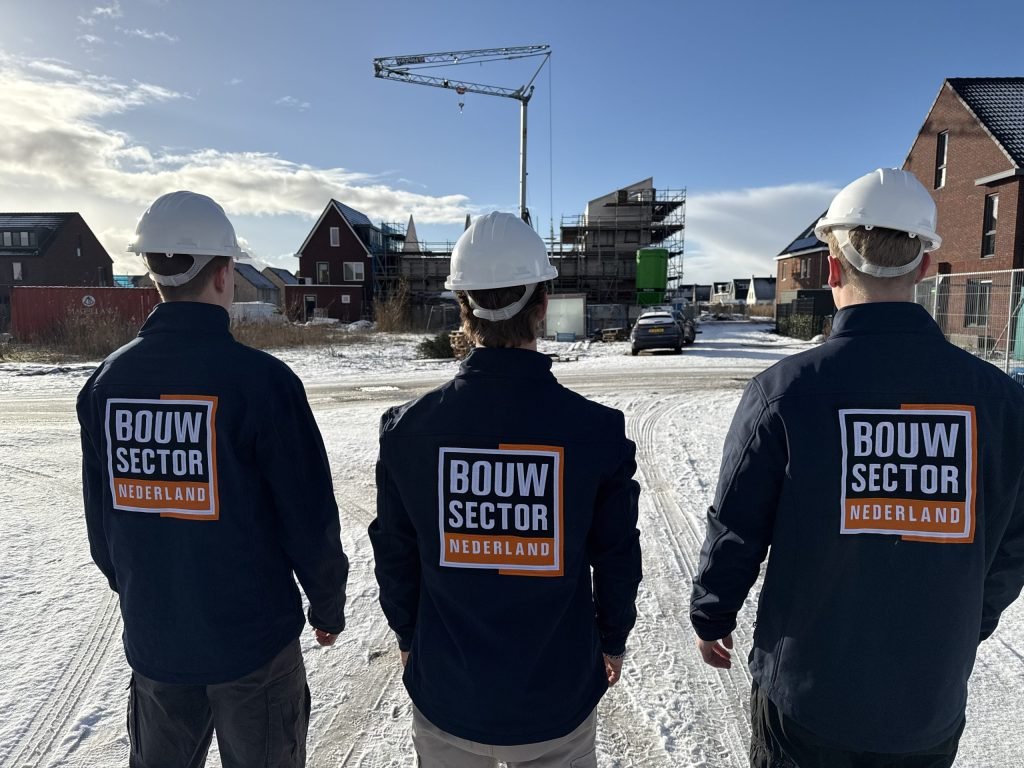 Drie vakmensen met witte helm en Bouwsector-jas op de rug lopen over een besneeuwde nieuwbouwlocatie met een bouwkraan